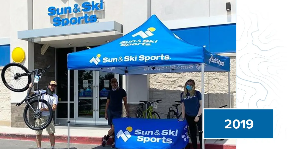A Sun and Ski Sports branded tent pitched outside of a Sun and Ski Sports store alongside three employees advertising bike services.