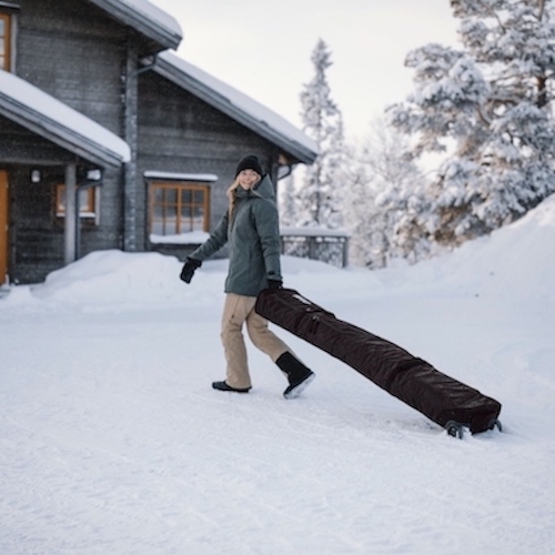 Woman traveling with skis and a snowboard using a ski bag from Sun & Ski Sports.