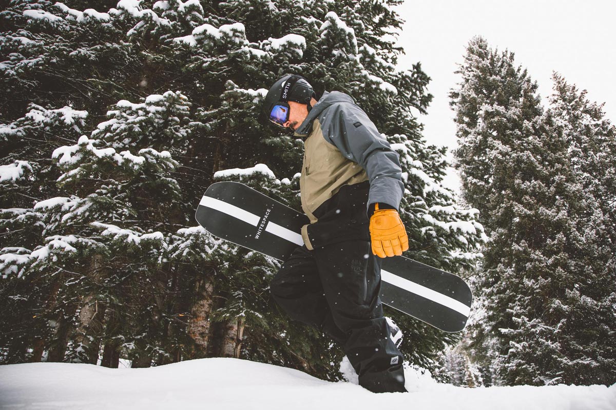  Man carrying a snowboard at his side while trudging through white snow next to tall snow-covered trees