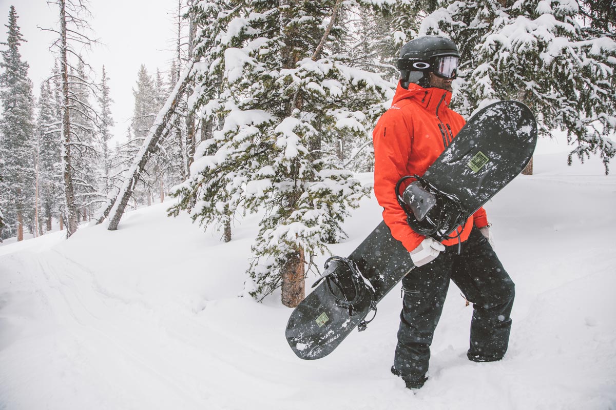 Snowboarder in a red puffer jacket carrying their black snowboard up a snow-covered mountain.