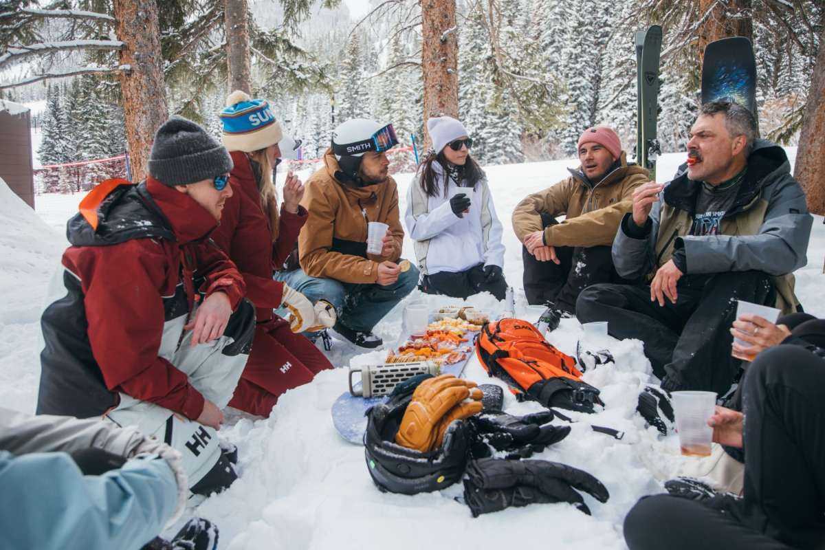 Group of friends dressed in ski gear hanging out and snacking in the snow