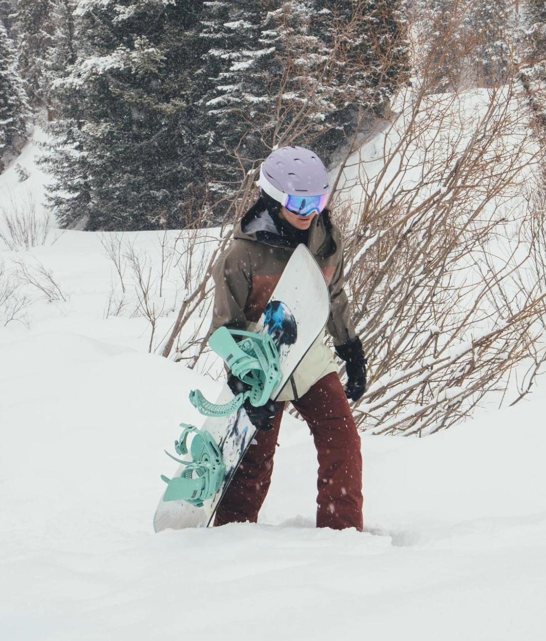 A snowboarder in a purple helmet and brown jacket wading through deep snow while carrying a white snowboard with mint green bindings.