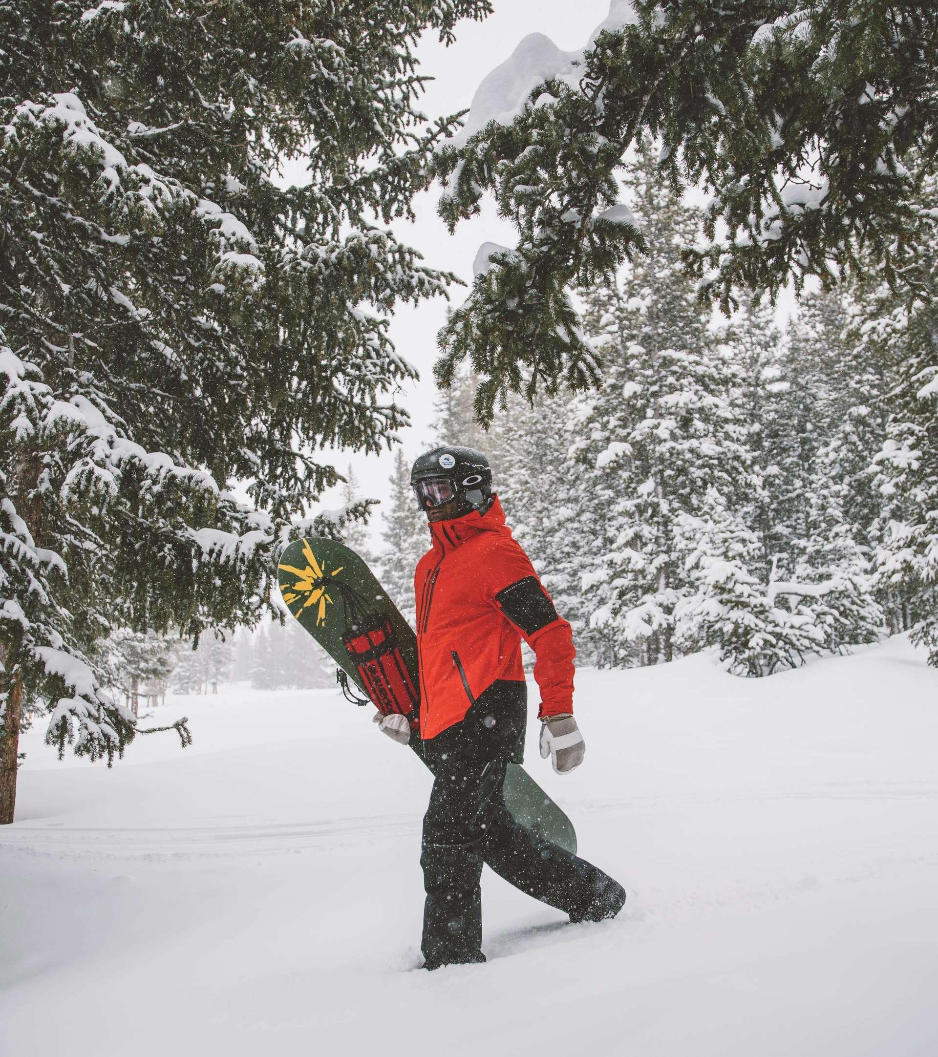 Man in a red and black snow jacket walking through the snow while carrying a board