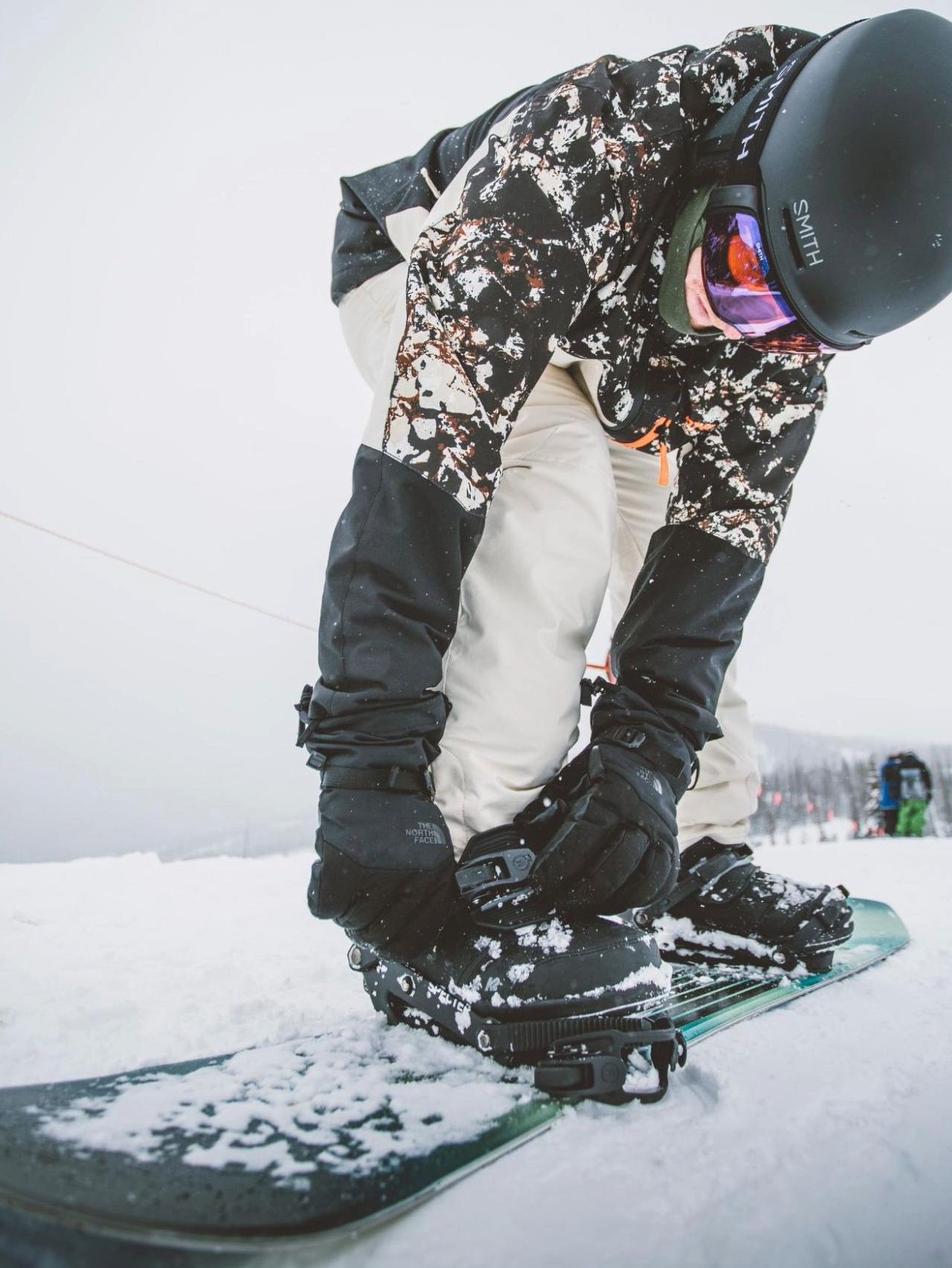 A person in a camouflage jacket and black helmet leaning over to strap their boots into snowboard bindings on a snowy slope.