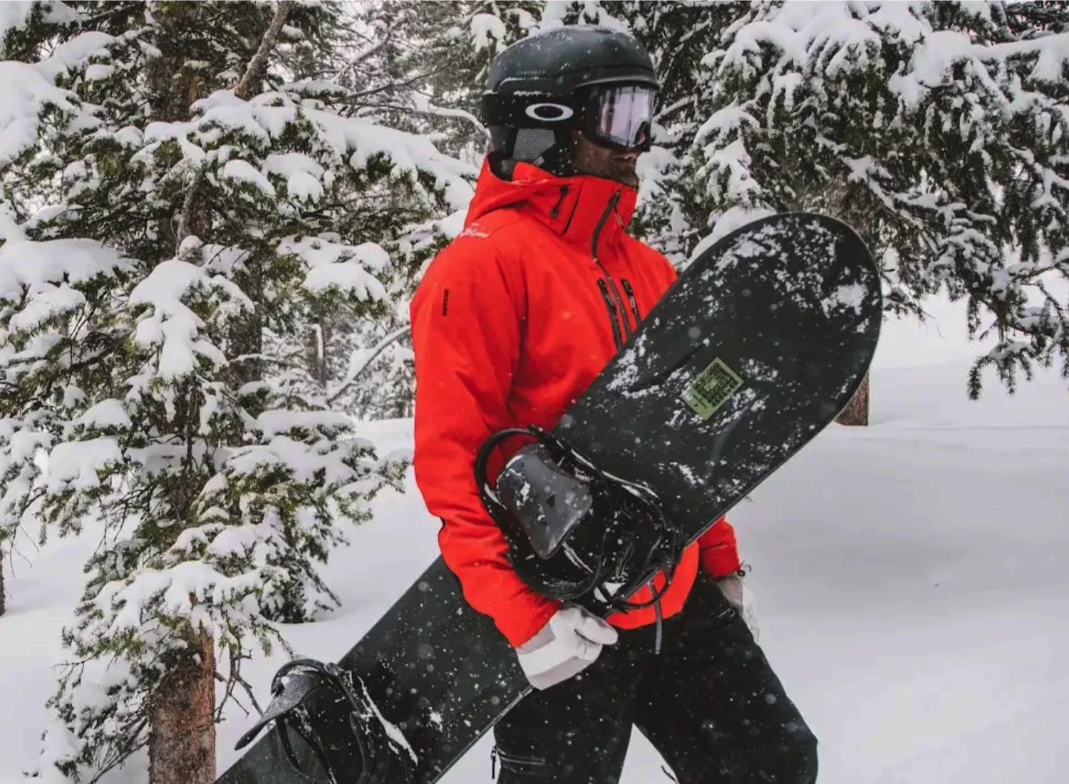 Man in a bright orange winter jacket holding a snowboard and walking through thick snow